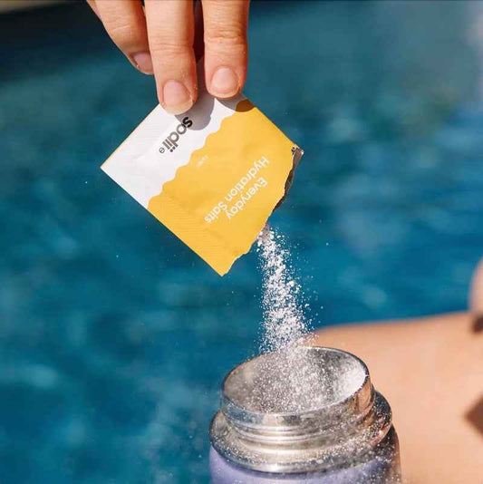 A hand pours a yellow and white Scopri Everyday Hydration Cube powder packet into a clear bottle, set against a blurred blue swimming pool background.