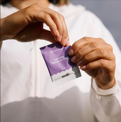 A close-up of a person's hands holding a purple and white sachet of sodii Everyday Hydration Swatches, with a white zip-up jacket in the background.