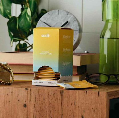 A box of sodii Everyday Hydration Gummies sits on a wooden table in a styled setting with books, a plant, glasses, and a green bottle in the background.